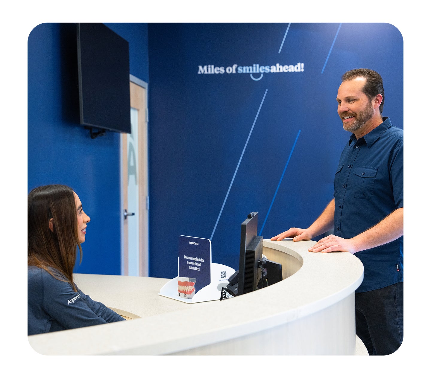 A smiling patient stands at the Aspen Dental front desk speaking with a team member, with a blue wall behind them displaying the phrase “Miles of smiles ahead!”