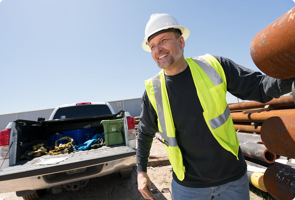 A male patient in a black long sleeve, a construction vest, and hard hat, smiling with new his dental implants as he stands in front of a truck, demonstrating how quickly ClearChoice patients are able to return to work with proper care and maintenance post-treatment.