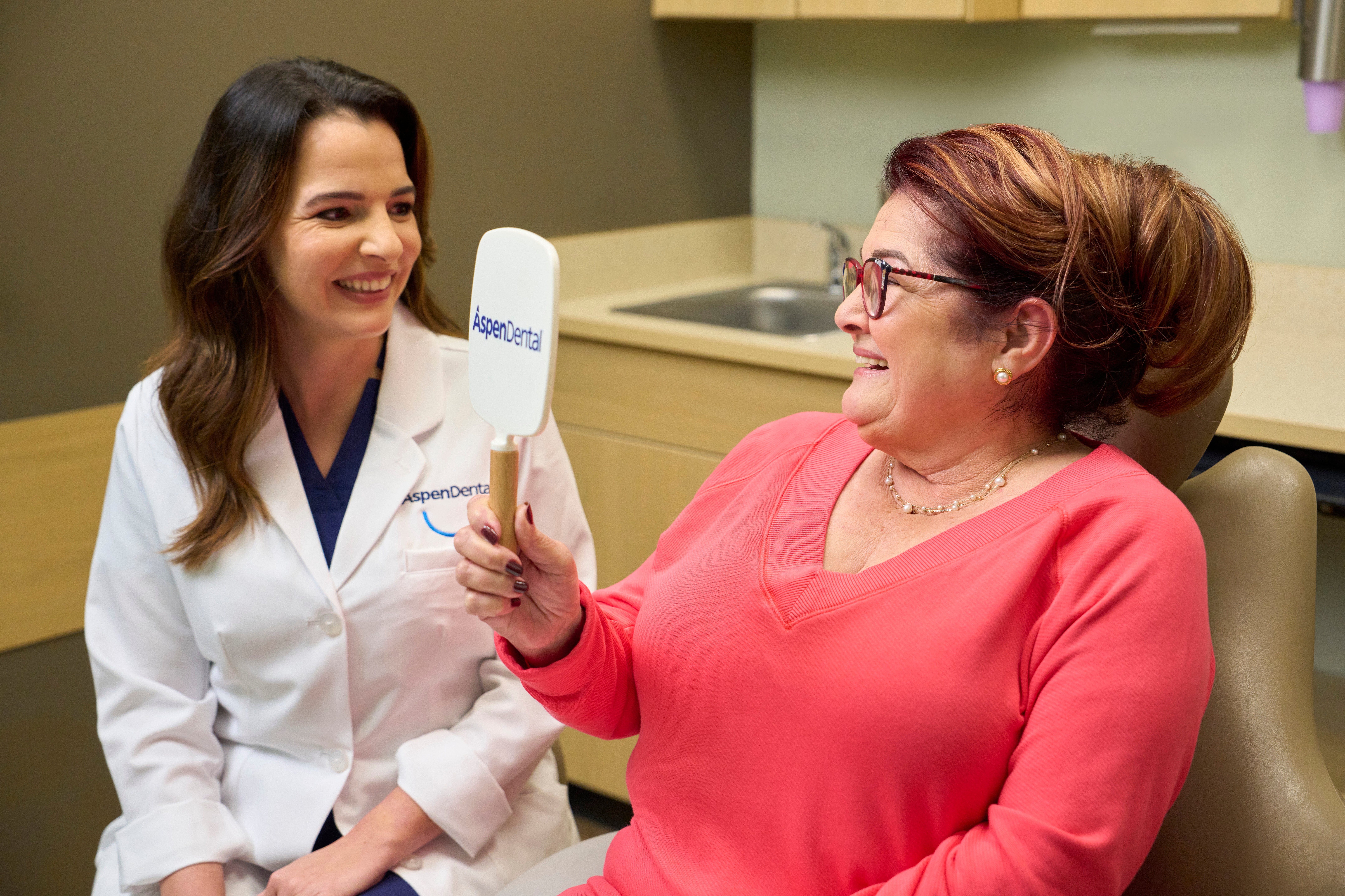 A woman is sitting in a dental chair looking at her teeth in a mirror, a dentist sits beside her