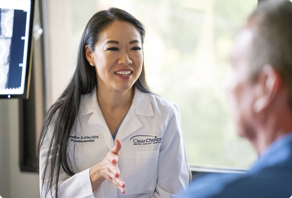 A ClearChoice prosthodontist in a white lab coat smiles at a senior patient seated across from her as she explains follow-up care and maintenance tips.