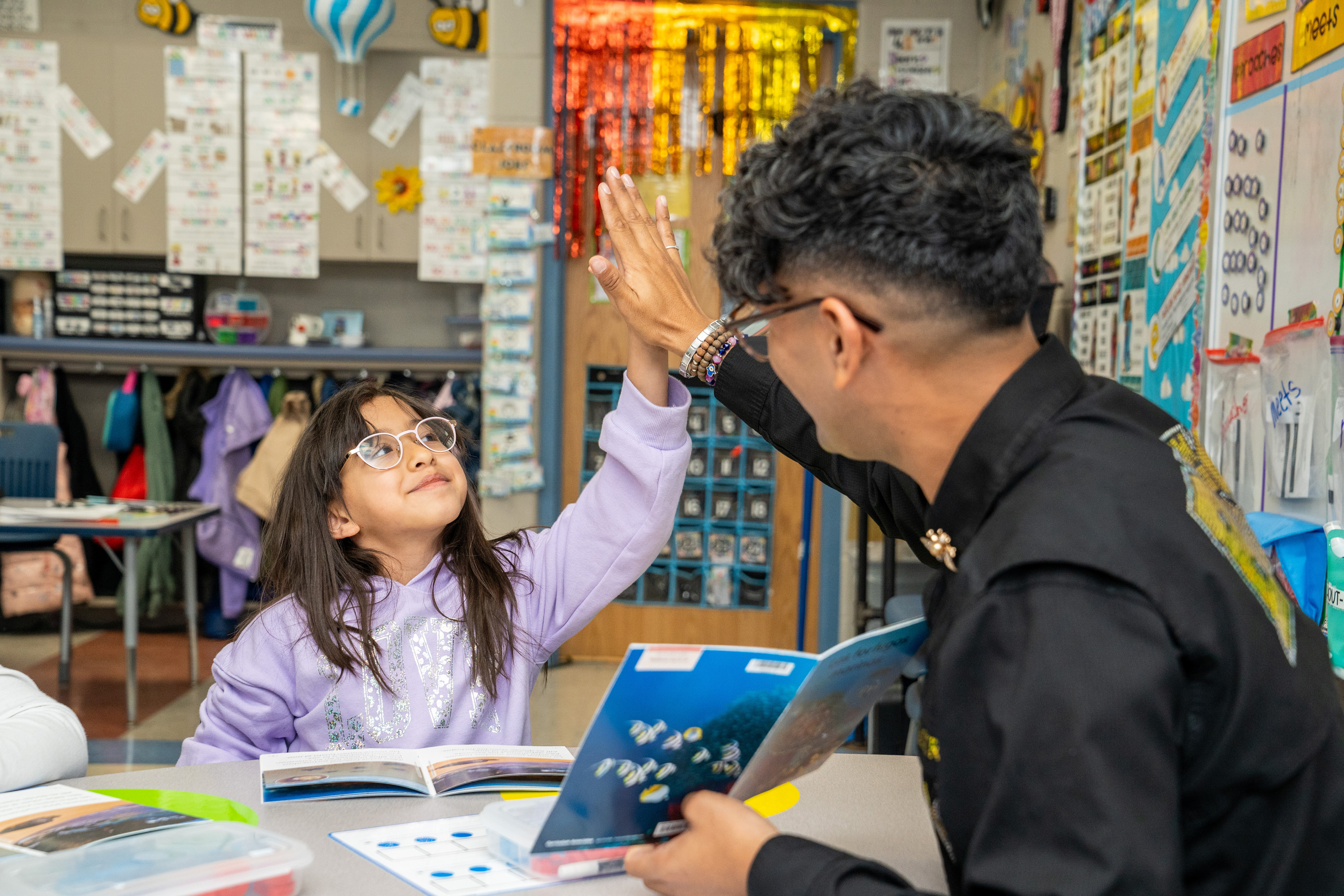 Teacher and a student sitting at a desk smiling and giving high fives with a book open in front of them