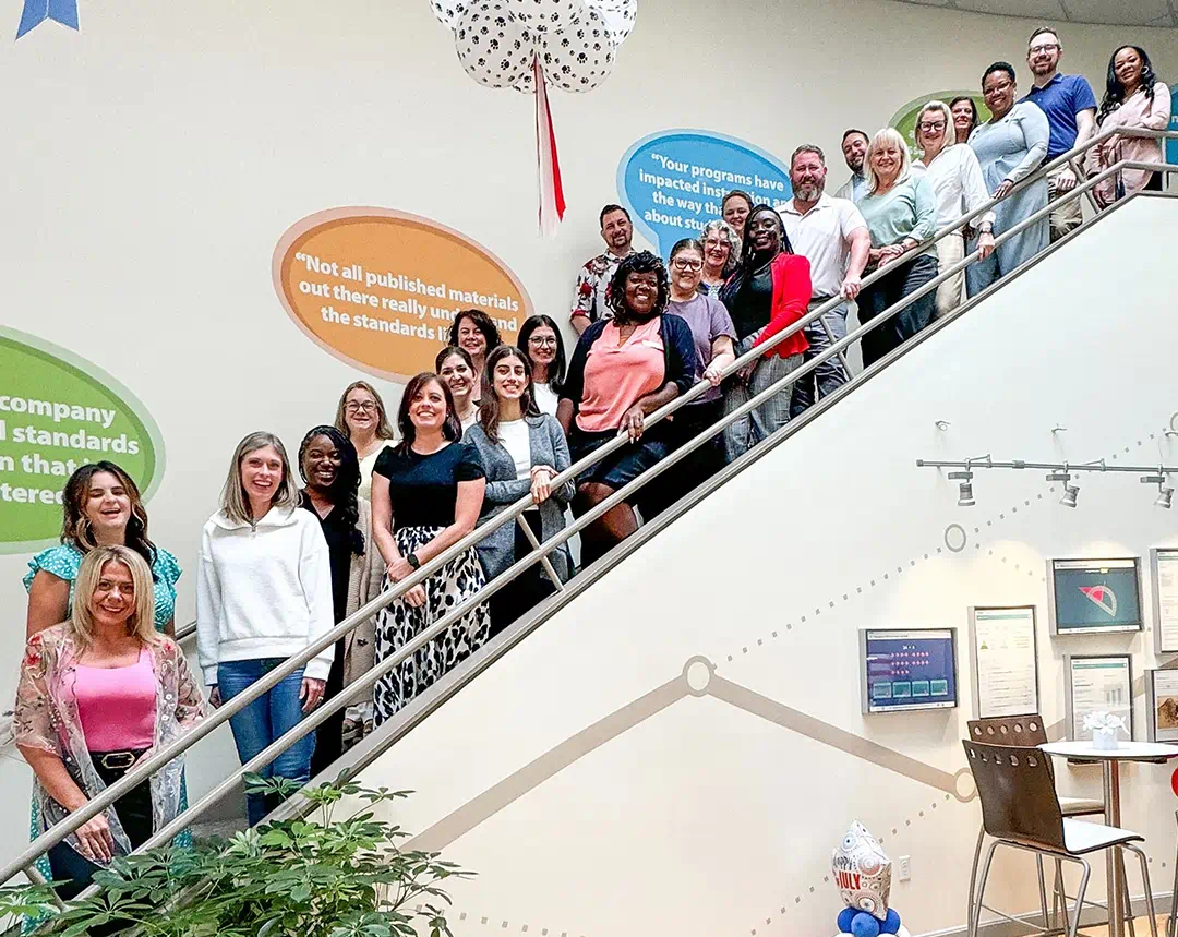 A diverse group of people poses on a staircase, smiling in front of colorful speech bubbles on the wall.