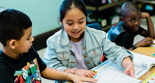 Three young students work together at a classroom table, smiling and engaging with an open textbook.