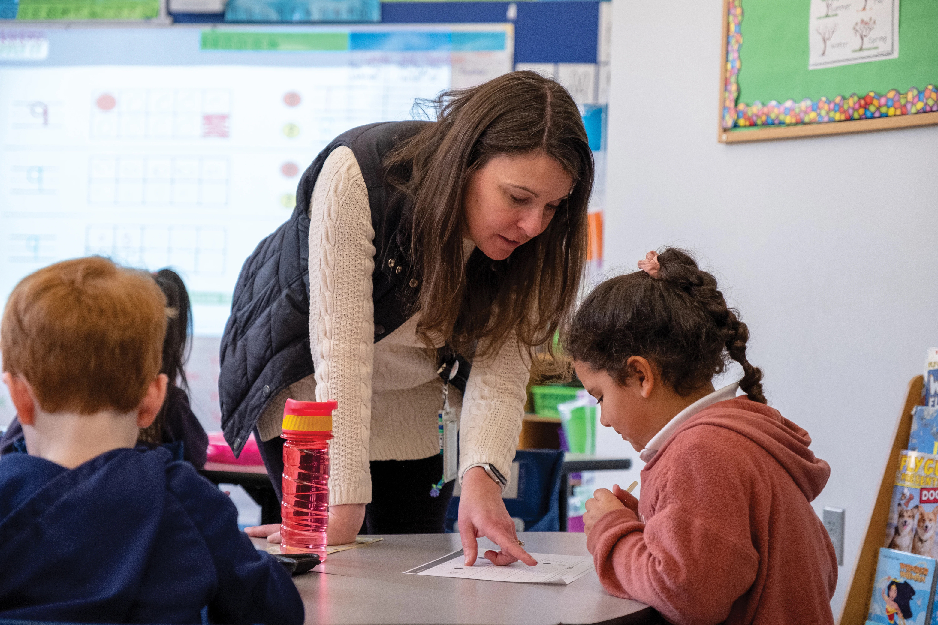 Teacher assists student at table, pointing to worksheet during classroom activity; other students work nearby.