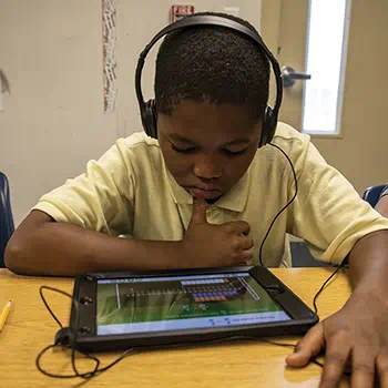 Young student wearing headphones, engaged with a learning activity on a tablet at a classroom desk.