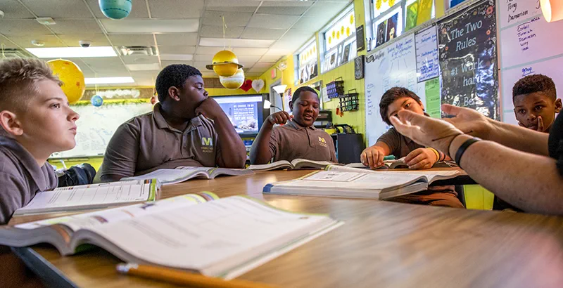 Middle school students sit around a table with open textbooks, engaged in a classroom discussion led by a teacher.
