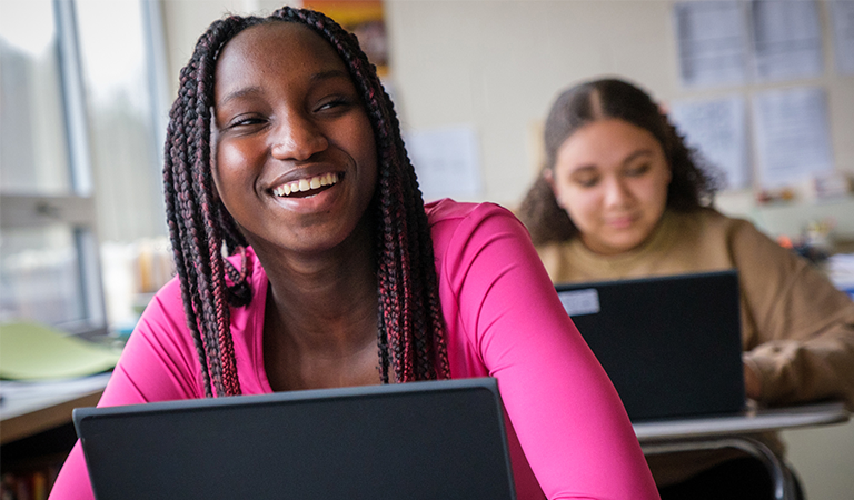 A smiling student at their desk with a laptop open and another student working in the background.
