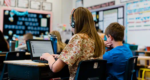 A student wearing headphones works on a laptop in a classroom while another student sits beside them, facing forward.