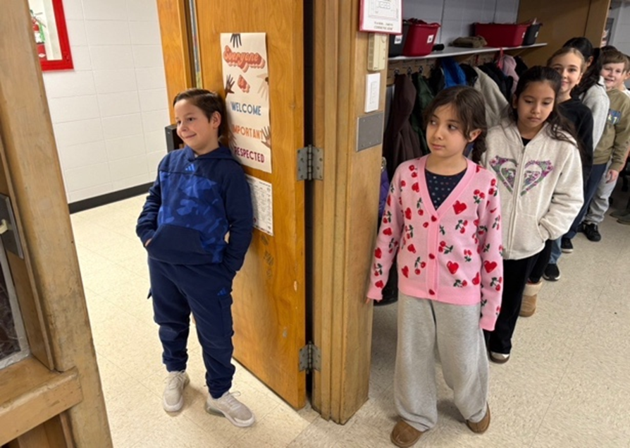 Elementary students stand in a line along a school hallway near a classroom doorway, with one student as line leader.