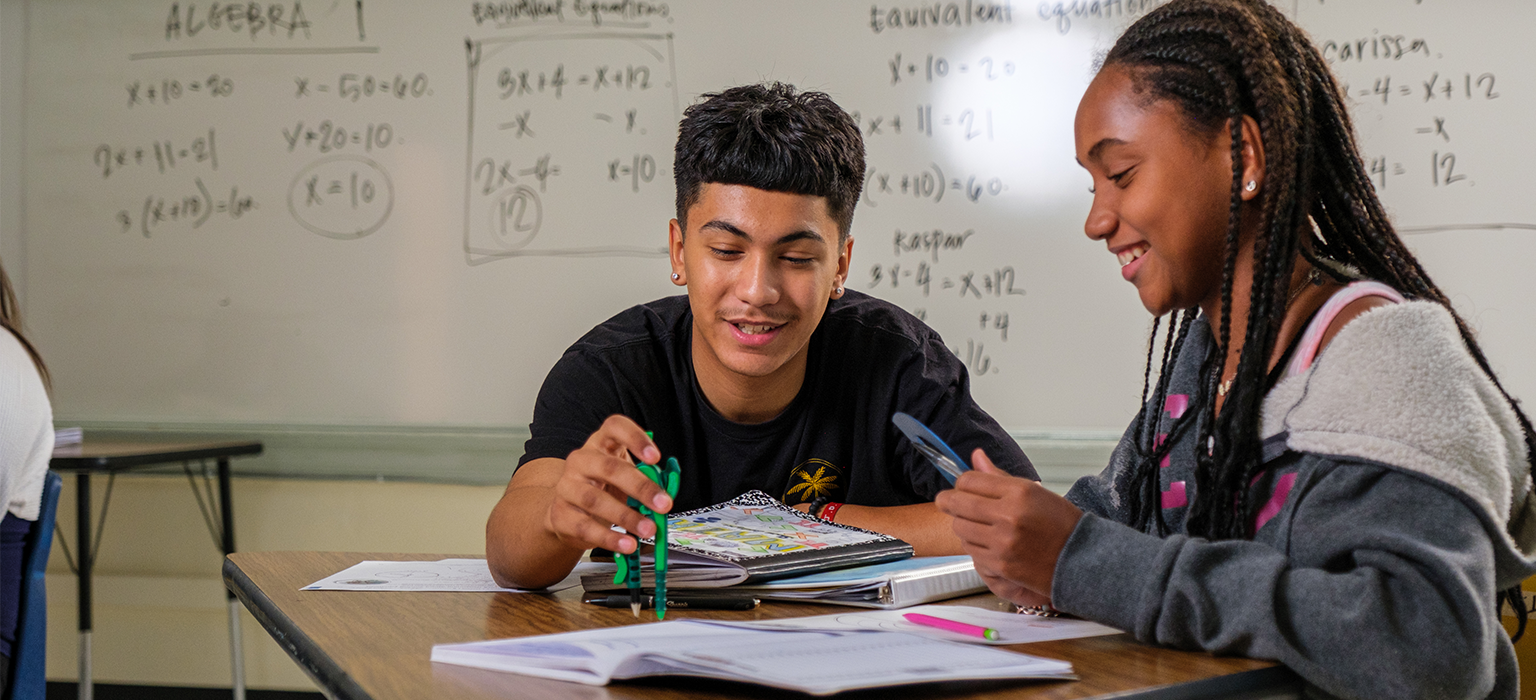 Students sit in front of a whiteboard filled with equations, partnering on a math assignment.