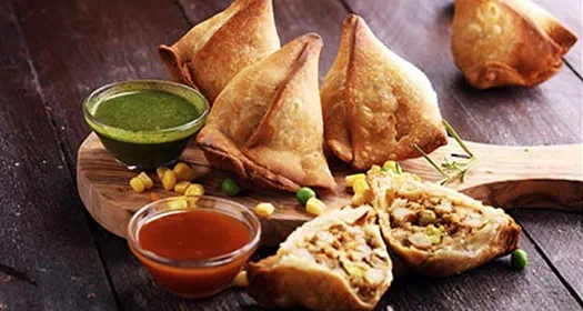 A wooden platter with crispy samosas, one cut open to reveal the filling, served with green and red dipping sauces, garnished with corn and peas.