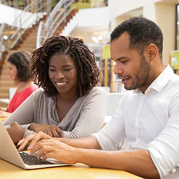 Two colleagues collaborate on a laptop in a modern library setting, with bookshelves and a staircase in the background.