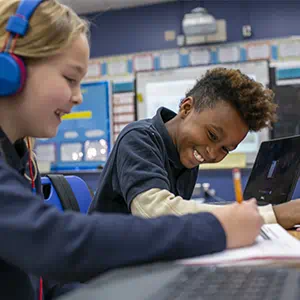 Two students in a classroom smiling while working on assignments using laptops and notebooks.