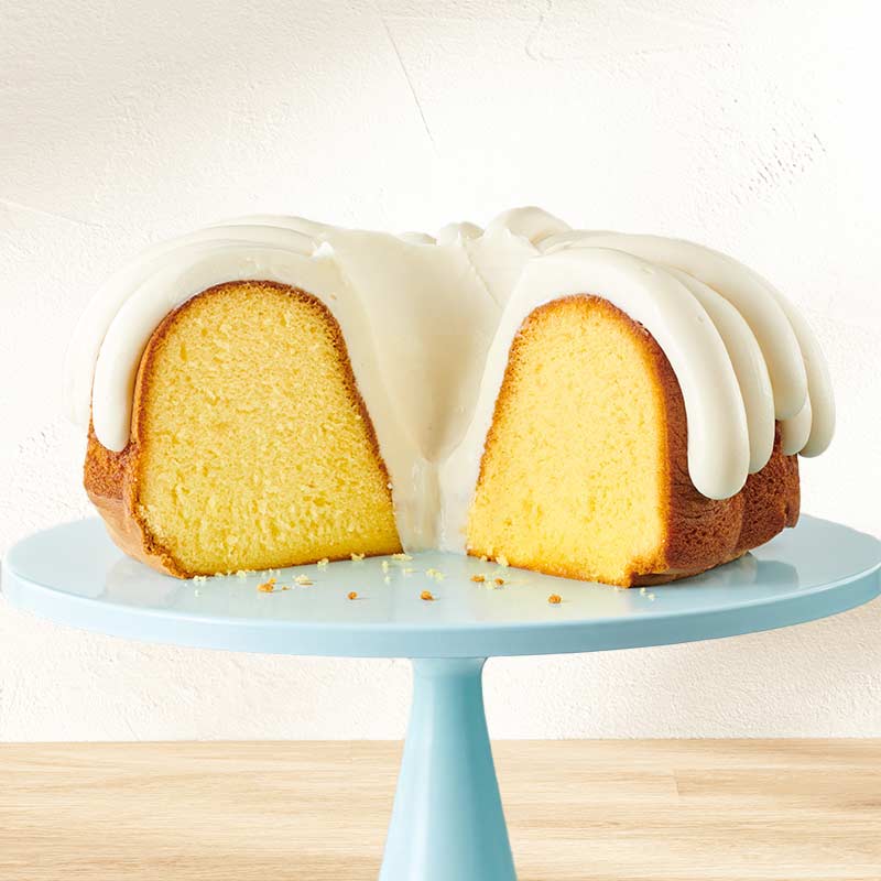 Lemon Bundt Cake with slice out of it on a cake stand, with signature frosting - Shop Bundt Cakes