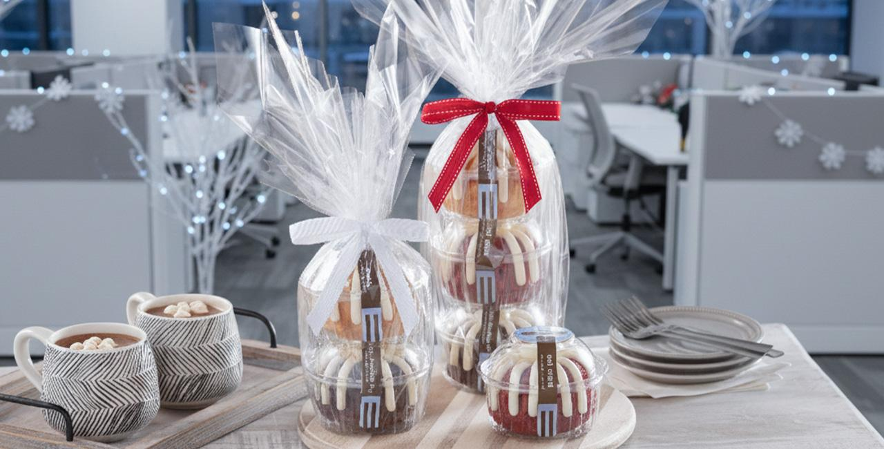 Gift wrapped Bundtlet towers packaged in clear cellophane with red and white ribbon bows, displayed on a table in an office setting with mugs and plates nearby.