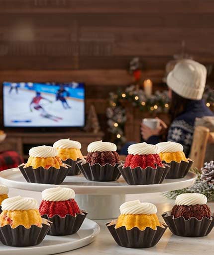 Bundtinis in assorted flavors topped with cream cheese frosting, displayed on tiered stands in a cozy winter setting with a TV showing a ski competition in the background.
