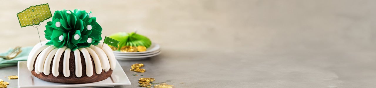 A frosted bundt cake topped with a green flower decoration and festive St. Patrick’s Day accents, displayed on a white platter with green and gold confetti.