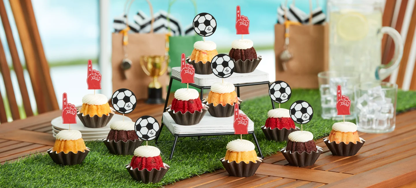 Mini Bundt cakes topped with cream cheese frosting and soccer and foam finger decorations, arranged on a sports themed dessert table for a game day celebration.