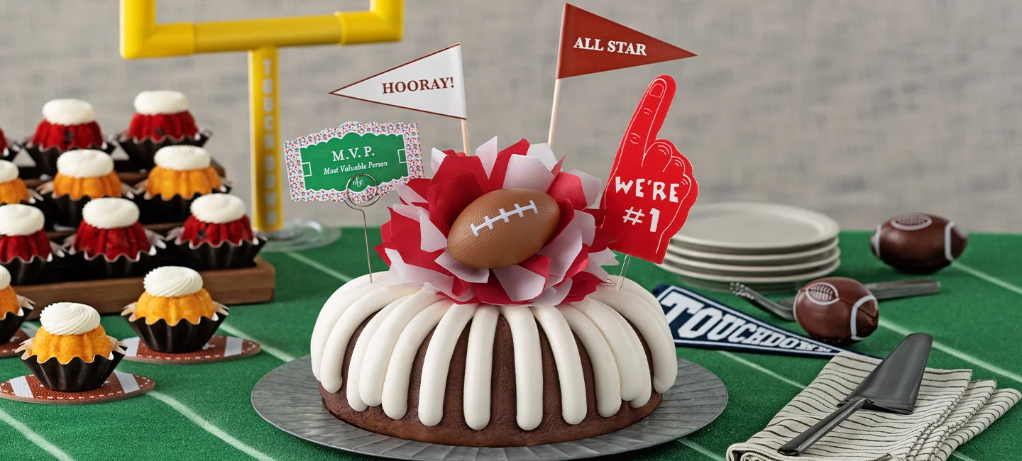 Bundt Cake topped with cream cheese frosting and a football decoration, and Bundtinis® on the side arranged on a sports themed dessert table for a game day celebration.