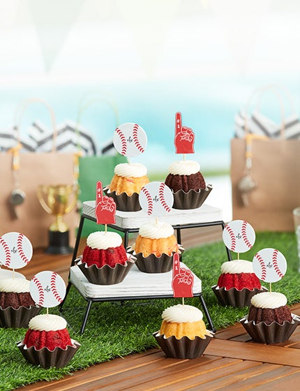 Bundtinis in assorted flavors topped with cream cheese frosting and adorned with baseball and foam finger decorations, displayed on a table for a sports themed celebration.