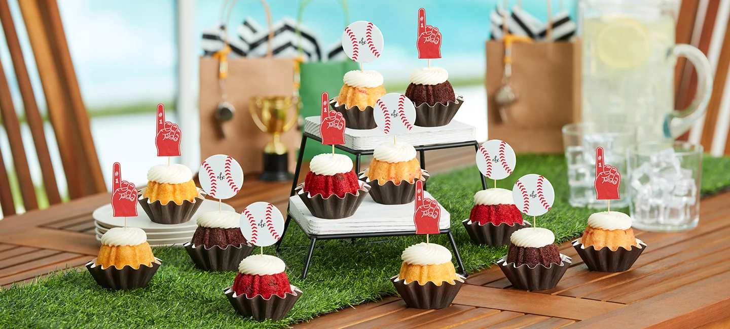 Bundtinis® topped with cream cheese frosting and baseball and foam finger decorations, arranged on a sports themed dessert table for a game day celebration.