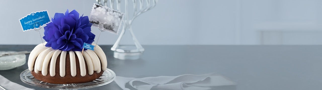 A bundt cake topped with signature frosting and a large blue bow, decorated with a &ldquo;Happy Hanukkah&rdquo; card, displayed on a glass cake stand with a menorah and festive silver accents in the background.