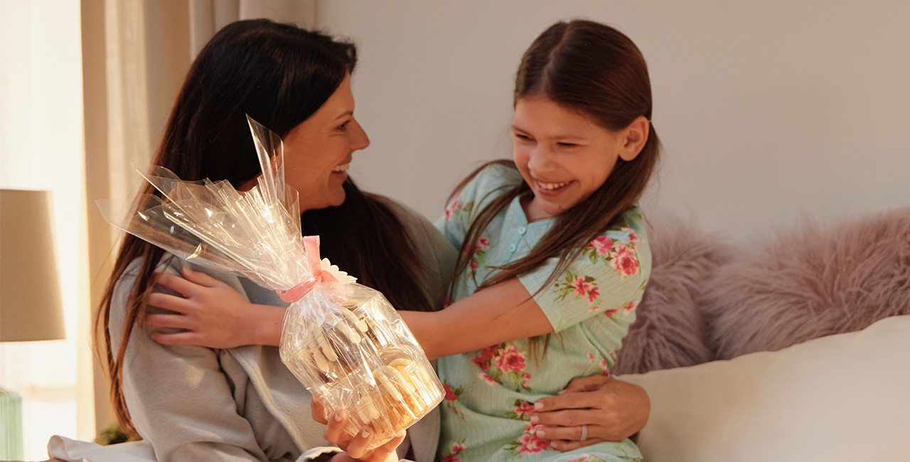 A woman and a smiling child share a hug while holding a gift wrapped Bundtlet tower tied with a pink ribbon in a cozy home setting.