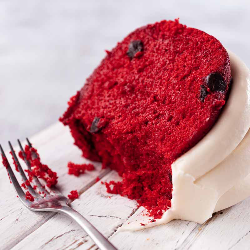 Red Velvet Bundt Cake with slice out of it on a cake stand, with signature frosting - Shop Bundt Cakes