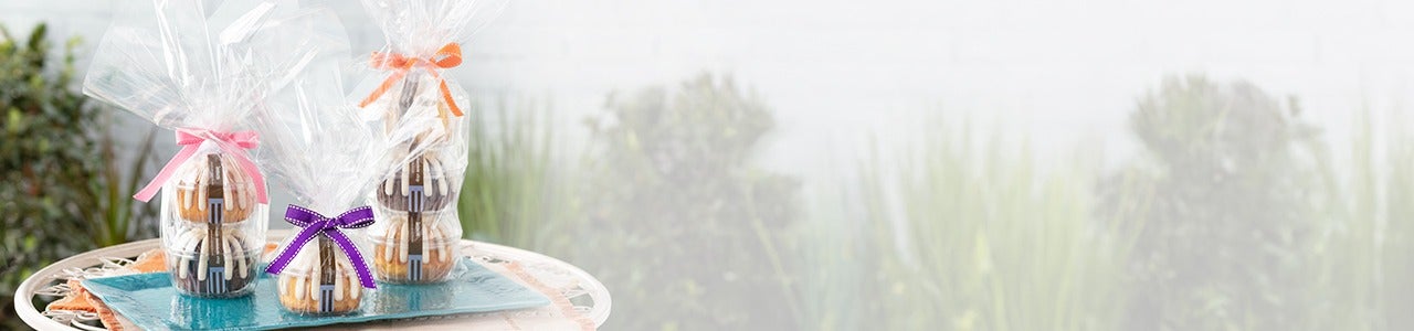 Three gift wrapped stacks of mini bundt cakes, each packaged in clear cellophane and tied with colorful ribbons pink, purple, and orange, displayed on a turquoise tray.