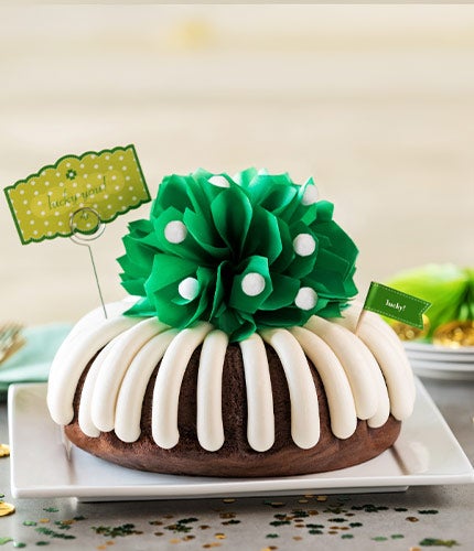 A frosted bundt cake topped with a green flower decoration and festive St. Patrick’s Day accents, displayed on a white platter with green and gold confetti.