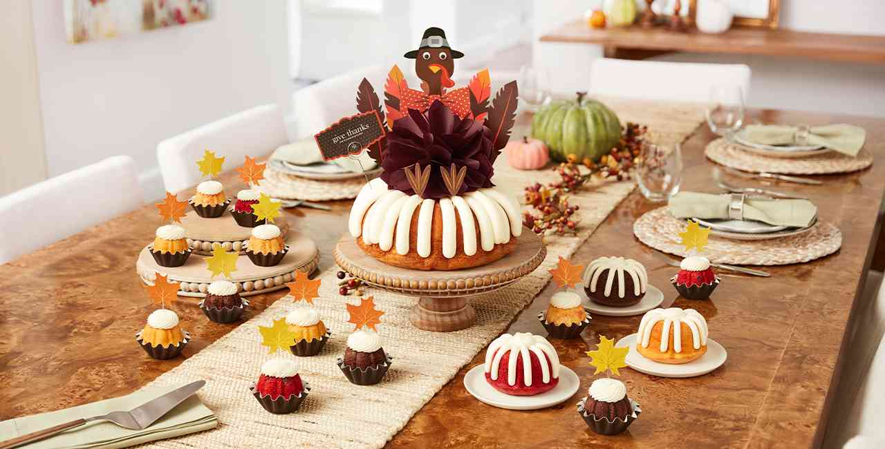 A large bundt cake with signature frosting and a turkey decoration on a cake stand and various bundtlets and bundtinis.