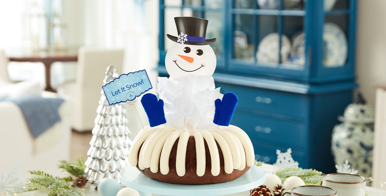 A frosted bundt cake topped with a cheerful snowman decoration, blue mittens, and a “Let It Snow!” sign, displayed on a cake stand with winter themed decor in the background.