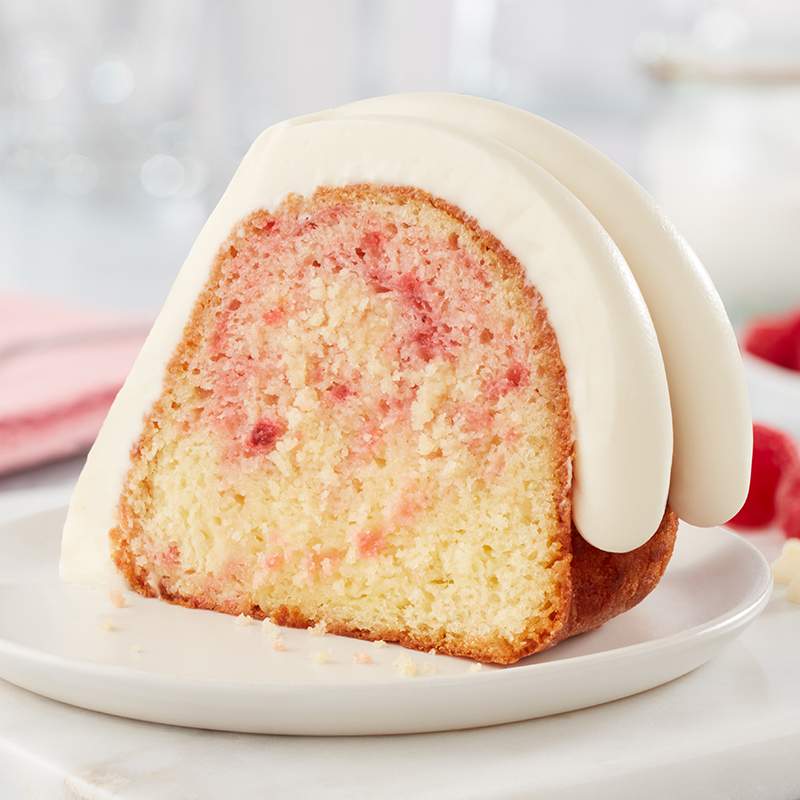 Strawberries & Cream Bundt Cake with slice out of it on a cake stand, showing chocolate chips baked into the cake and signature frosting on top - Shop Bundt Cakes