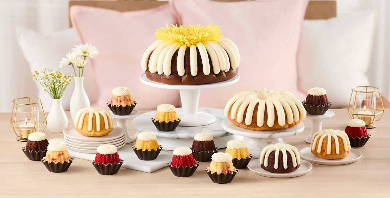 Assorted bundt cakes including classic large bundt cakes, bundtlet, and bundtinis topped with cream cheese frosting, arranged on cake stands for a celebration dessert display.