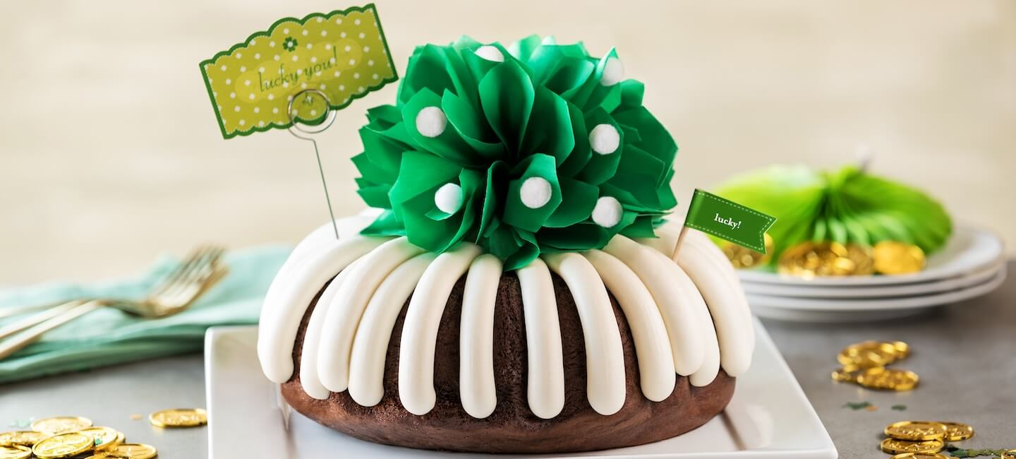 A bundt cake topped with cream cheese frosting and a green decorative bow, styled for a St. Patrick’s Day celebration with gold coins on the table.