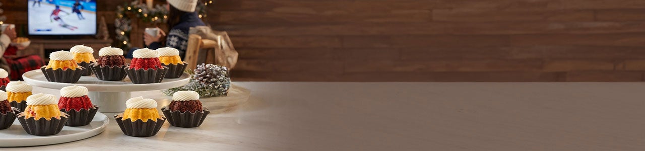 Bundtinis in assorted flavors topped with cream cheese frosting, displayed on tiered stands in a cozy winter setting with a TV showing a ski competition in the background.