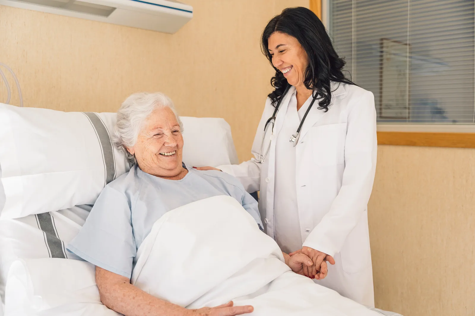 A doctor smiling and holding the hand of a senior patient in a hospital bed.