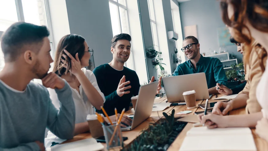 A group of people are sitting around a table with laptops
