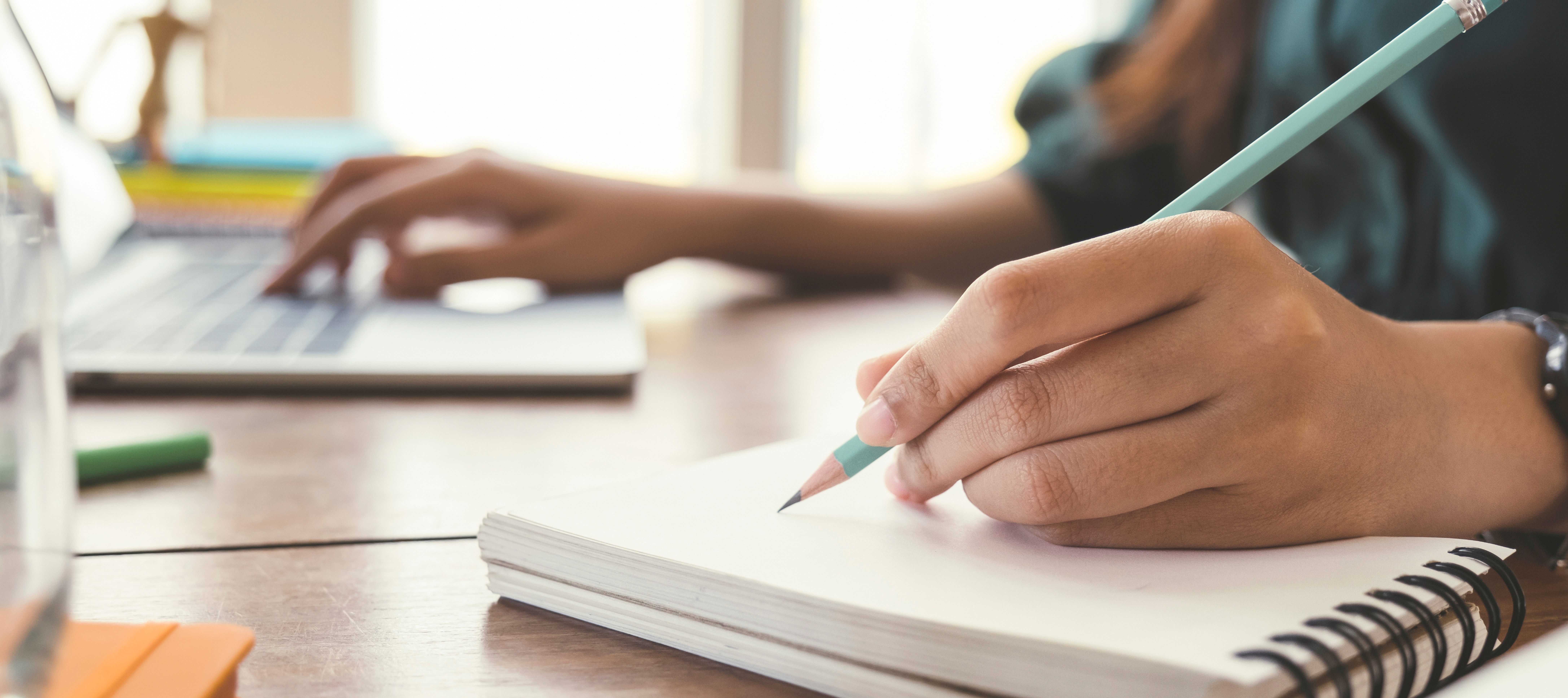 A person is writing in a notebook while using a laptop