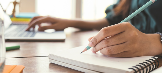A person is writing in a notebook while using a laptop
