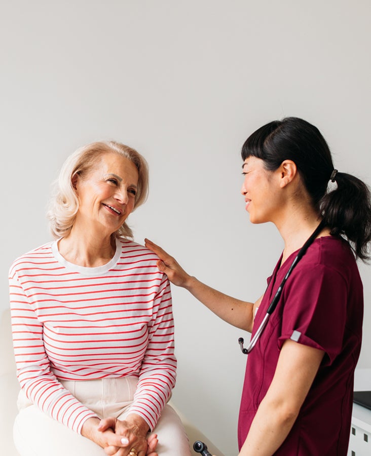 A healthcare professional in a maroon uniform talks to a smiling older woman in a striped shirt, with a stethoscope around the professional's neck.