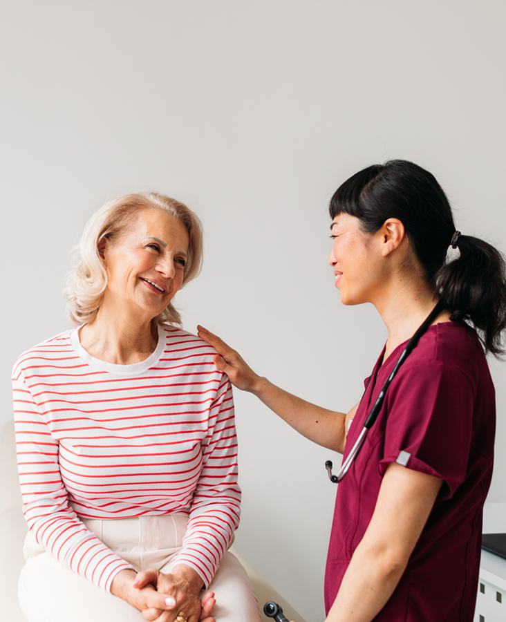 A healthcare professional in a maroon uniform talks to a smiling older woman in a striped shirt, with a stethoscope around the professional's neck.