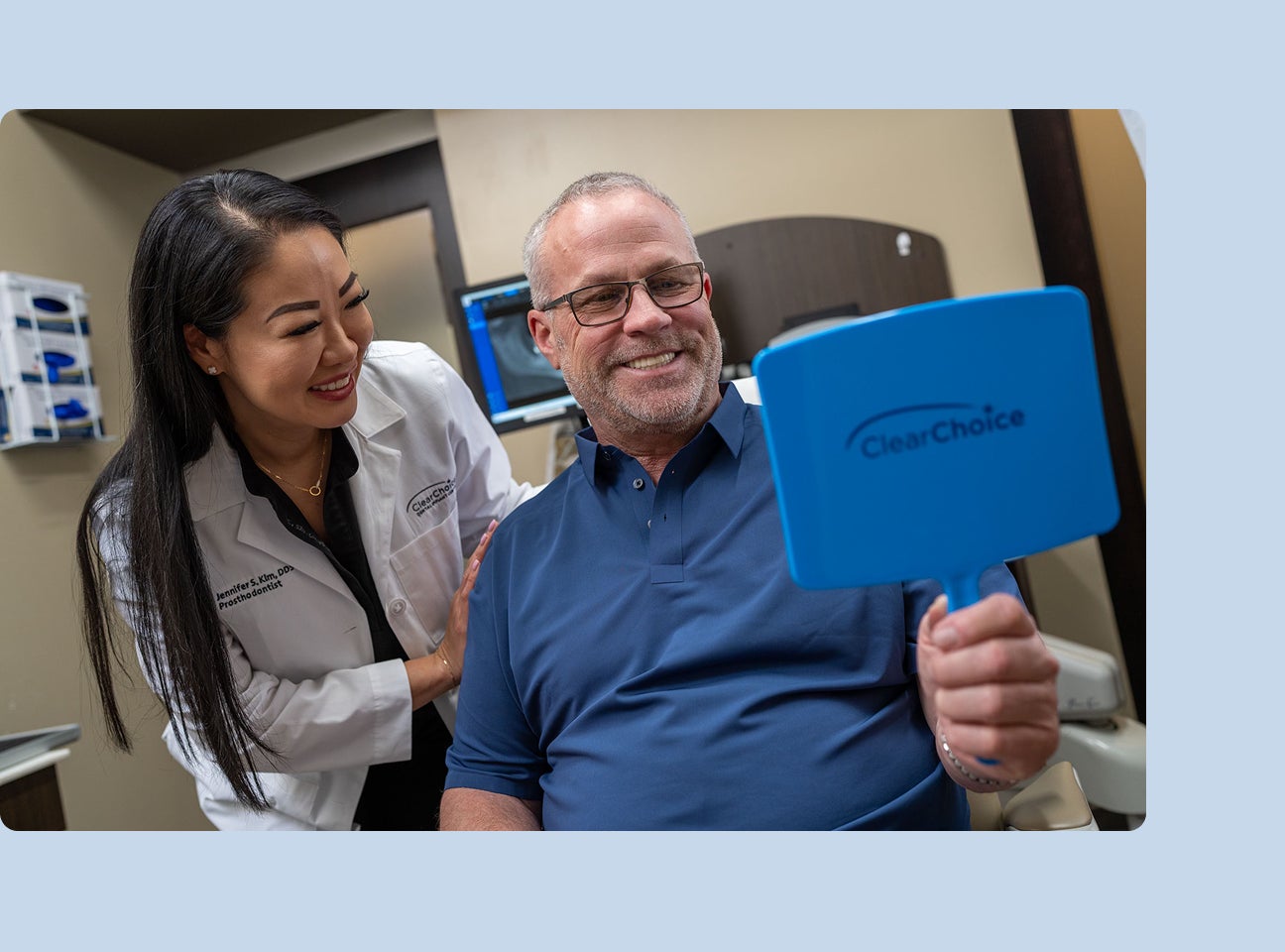 An image of a ClearChoice patient in a blue shirt admiring his new set of dental implants in a hand-held mirror with his doctor standing beside him.