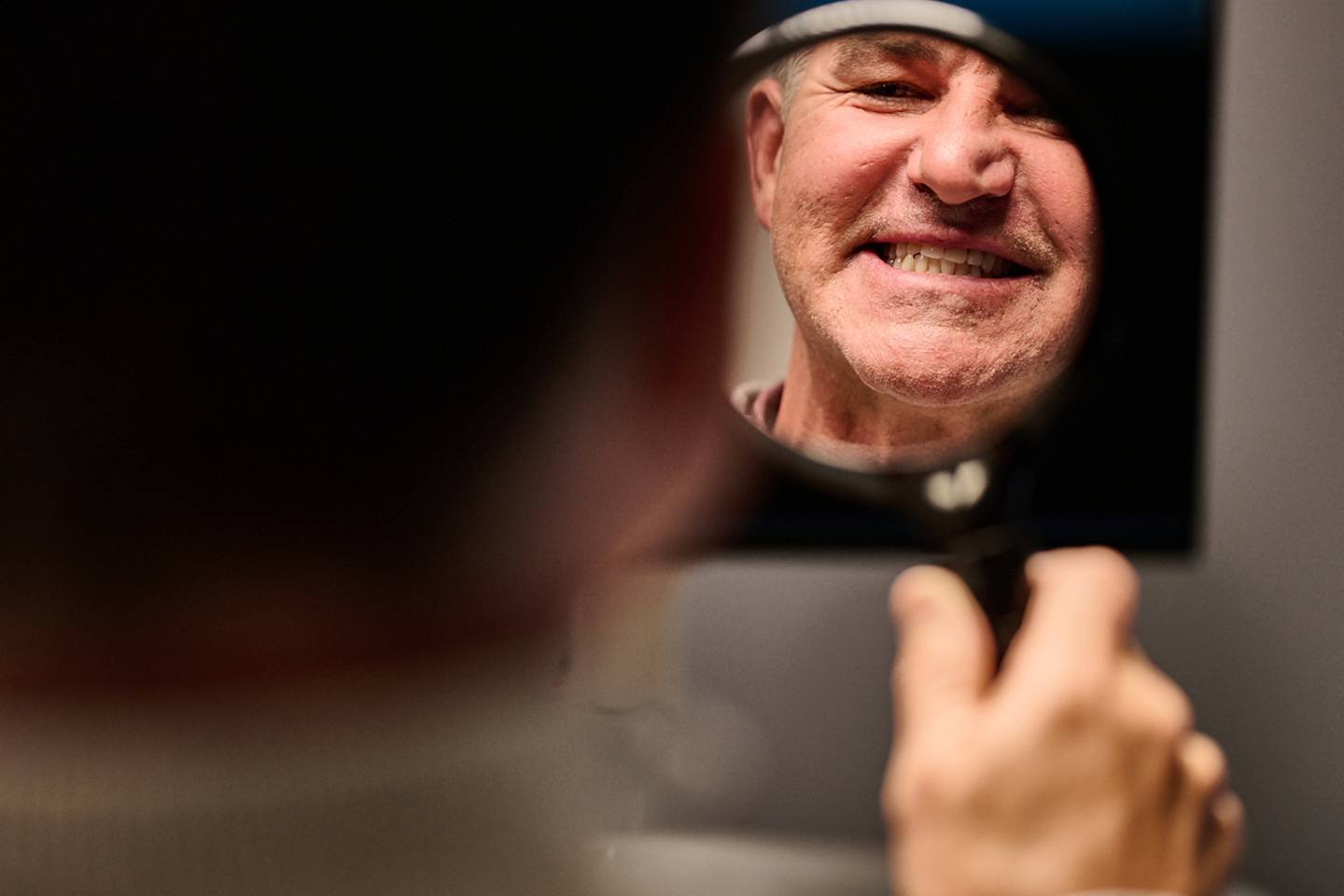 A man smiling in the mirror while checking his dentures, reflecting confidence and satisfaction. Aspen Dental supports patients in achieving a natural and comfortable smile with dentures.