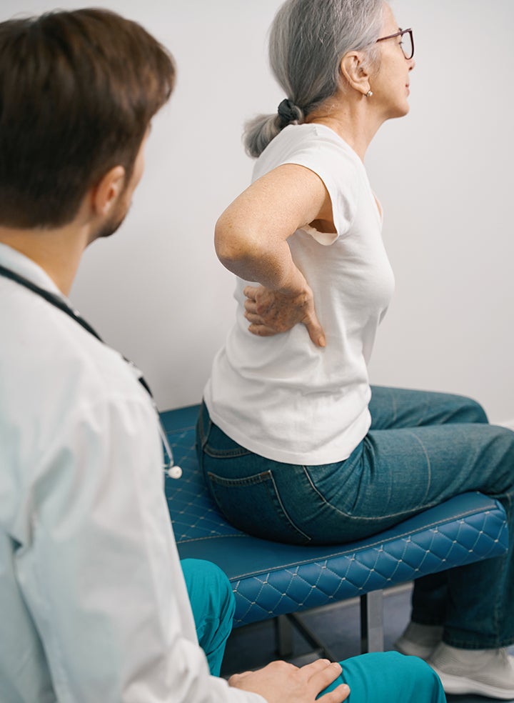 An elderly woman is sitting on a bed holding her back while a doctor examines her