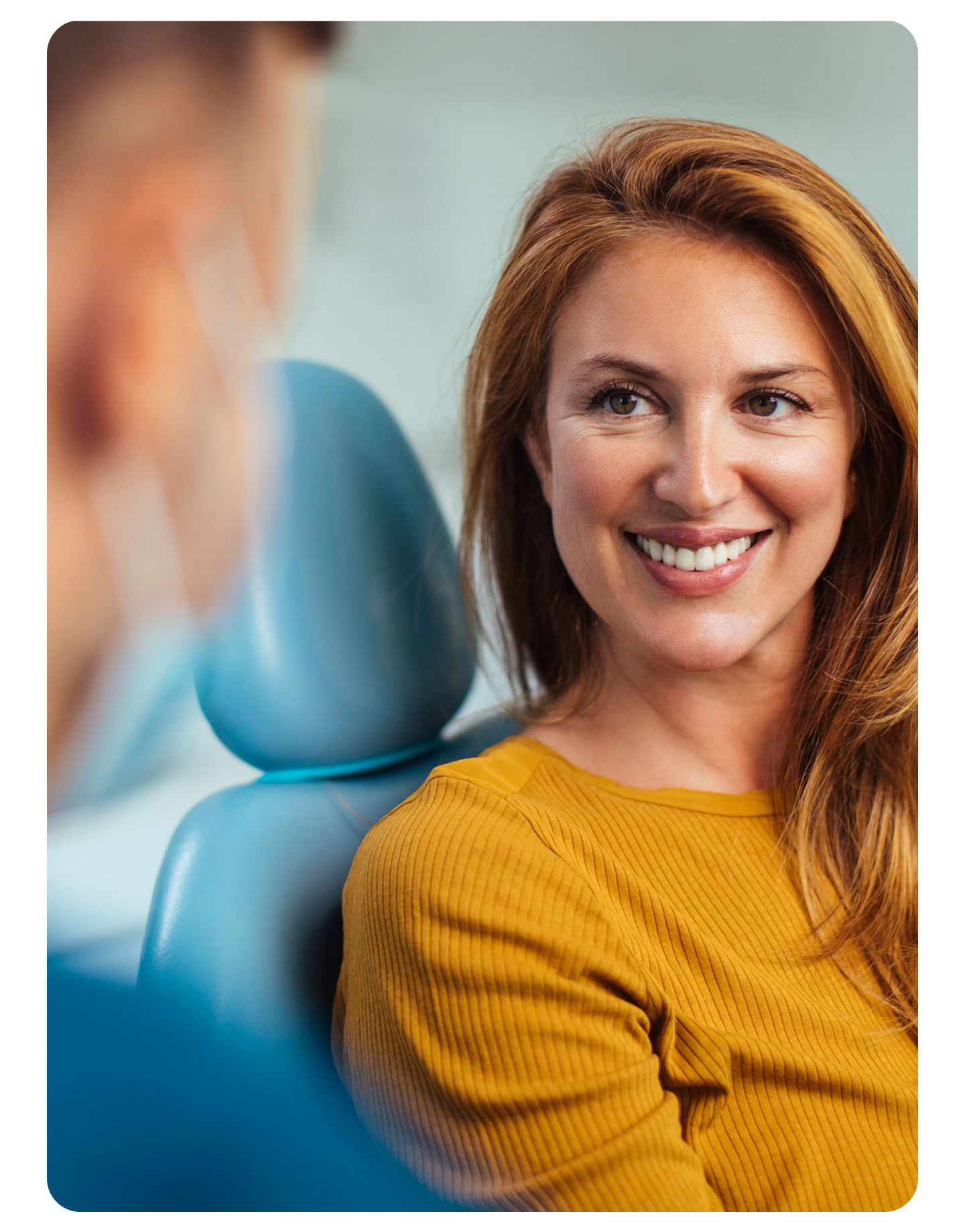 A woman in a mustard sweater smiles while speaking with a masked dental professional, seated in a dental chair during a consultation about bad breath.