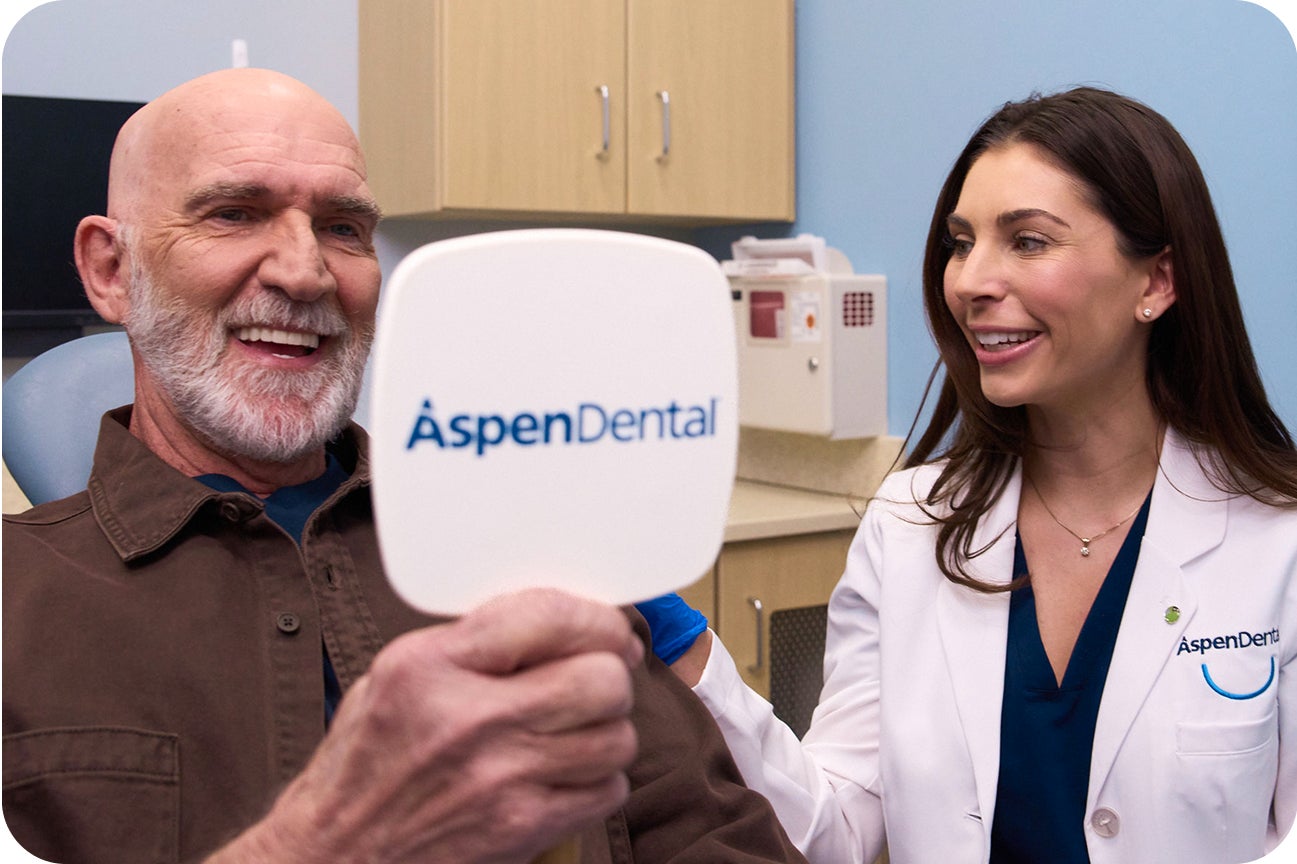 A happy Aspen Dental patient admires his smile in a handheld mirror after receiving snap-in dentures as a smiling Aspen Dental dentist stands next to him with her hand on the patients shoulder. The image conveys confidence symbolizing the benefits of snap-in dentures.