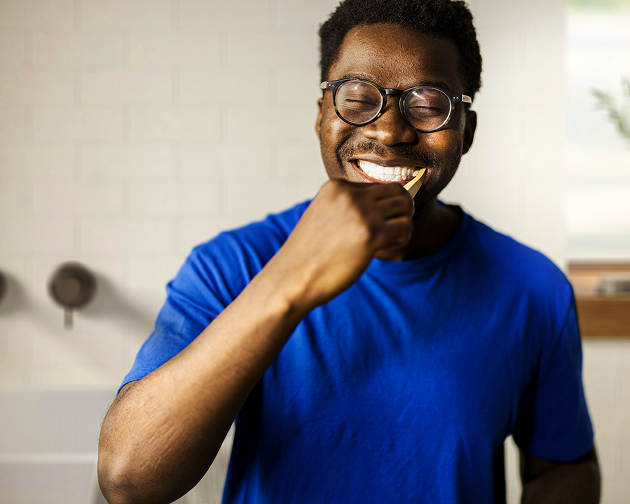 A smiling man in a blue shirt brushes his teeth in front of a mirror, emphasizing good oral hygiene habits. Brushing regularly helps prevent cavities and gum disease. Aspen Dental promotes healthy dental care routines for strong teeth and gums.