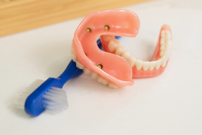 Close-up of snap-in dentures with visible implant attachments placed next to a blue cleaning brush, illustrating proper denture care, Aspen Dental branding.