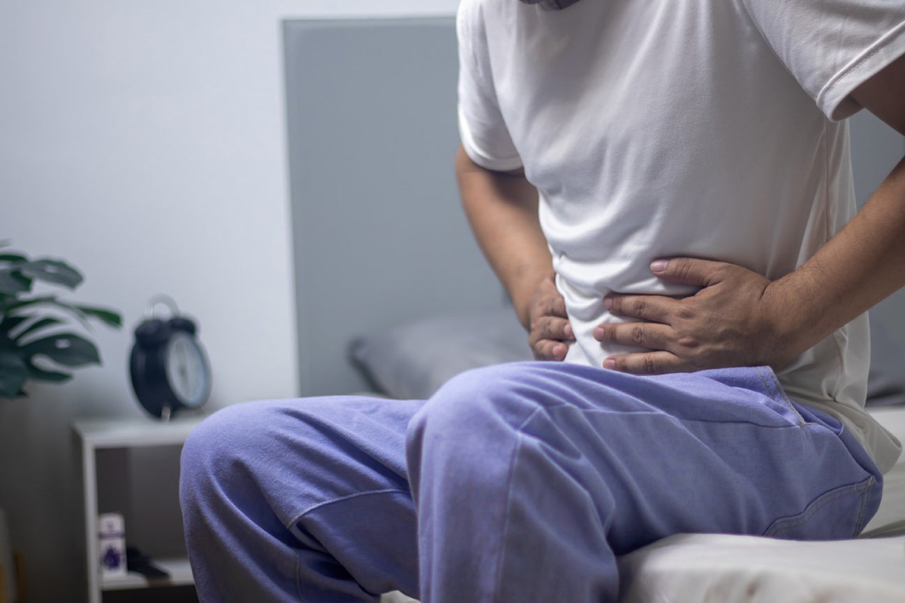 A zoomed in photo of the middle of a person's body, they are sitting on a bed and clutching their stomach in pain.